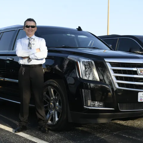 Man smiling in sunglasses beside a black Cadillac SUV in a parking lot.