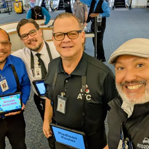 Four smiling men in uniforms at an airport holding electronic tablets.