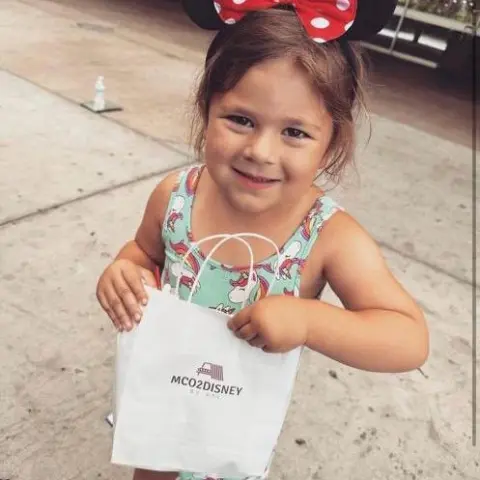 Child with Minnie Mouse ears holds a small MCO2Disney bag at an airport.