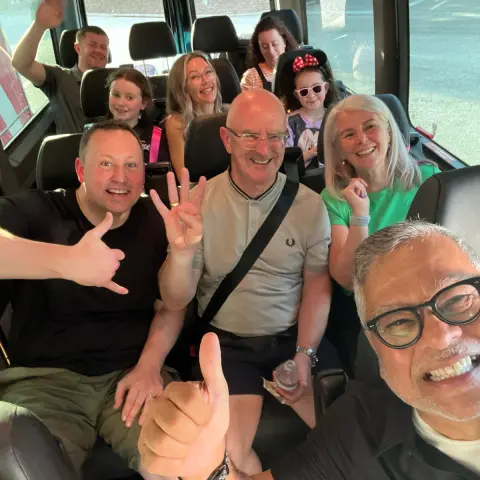Group of smiling people sitting in a van, waving and giving thumbs up.