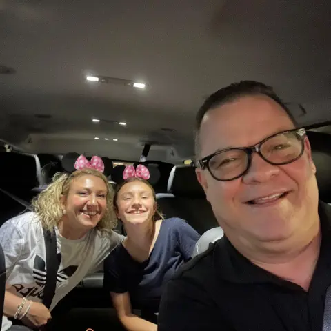 Three people smiling inside a car, one wearing pink mouse ears.