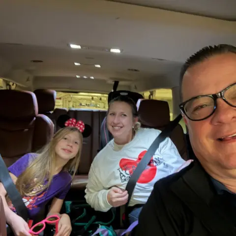 Three people smiling inside a car, two wearing seatbelts, including a child with Minnie Mouse ears.