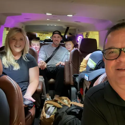 Smiling family inside a car with brown leather seats, bags visible.