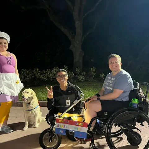 Three people, a dog, and two wheelchairs in a park at night, with one person in costume and a sign on one wheelchair.