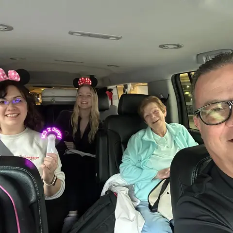 Four people in a car smiling, two wearing Minnie Mouse ears, with luggage in the back.