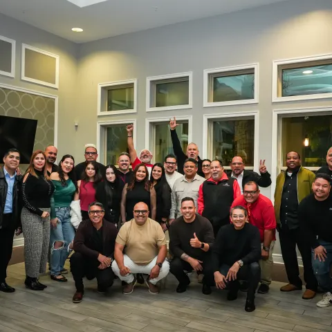 Group of 24 people posing indoors, some raising hands, with large windows in the background.