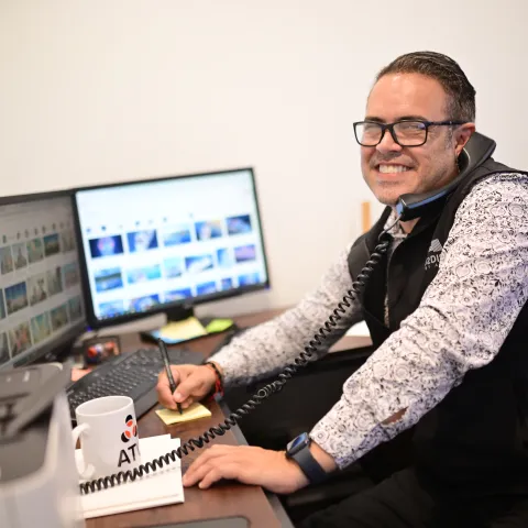 Person smiling while multitasking at a desk, using a phone and computer.